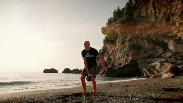 A man performs kettlebell exercises on a picturesque beach at sunrise.