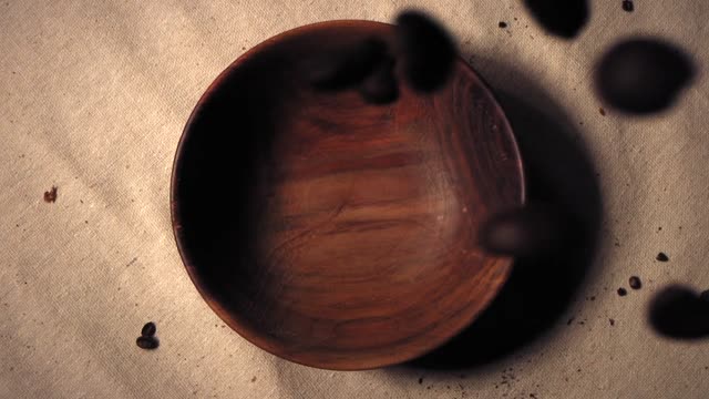 Aromatic coffee beans pour into a rustic wooden bowl on a textured surface.
