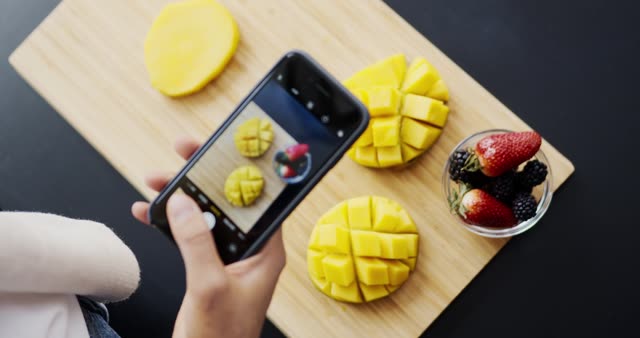 Person takes artistic shots of mangoes and berries on wooden board.