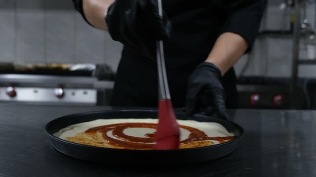 Chef preparing pizza with sauce in a commercial kitchen setting.