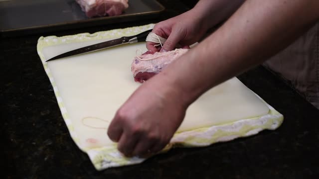 Close-up of hands tying raw meat on a chopping board with knife.