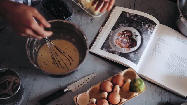 Hands mixing pancake batter with a whisk by an open cookbook on a kitchen table.