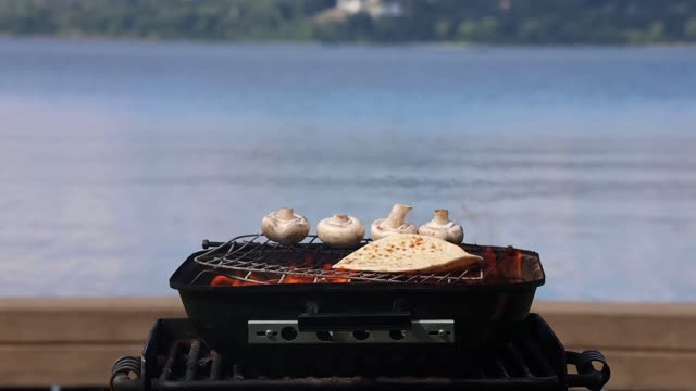 Outdoor grilling of naan and mushrooms with a lake view.