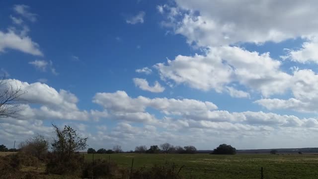 Time-lapse of cumulus clouds drifting over a green field under a blue sky.